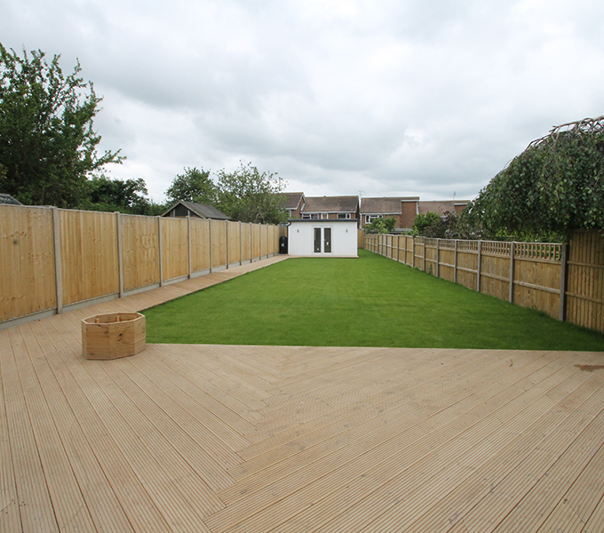 a long garden with a wooden decking and lawn with a white building at the bottom of the garden